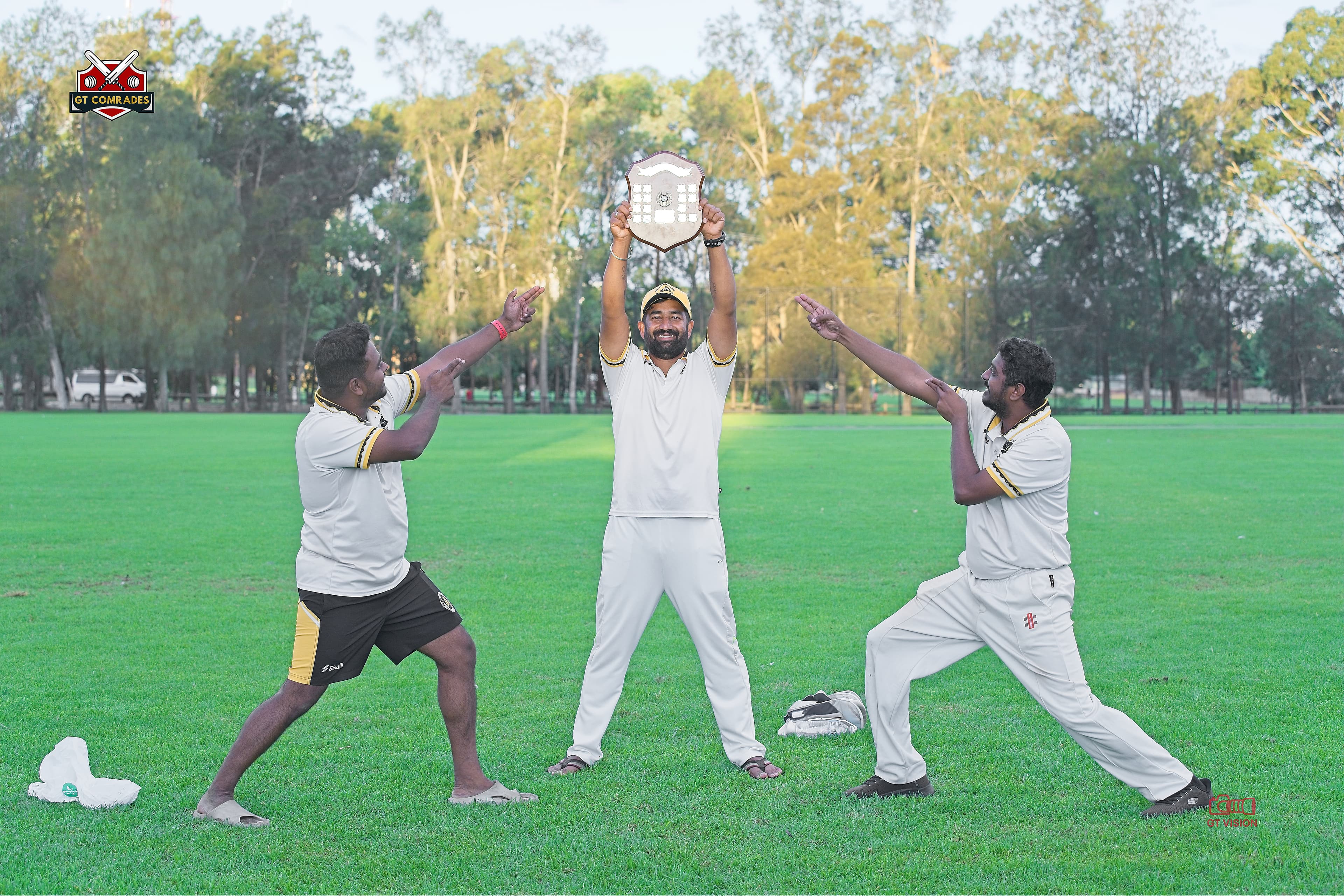 Three GT Comrades players celebrate as one raises a championship shield on the cricket field